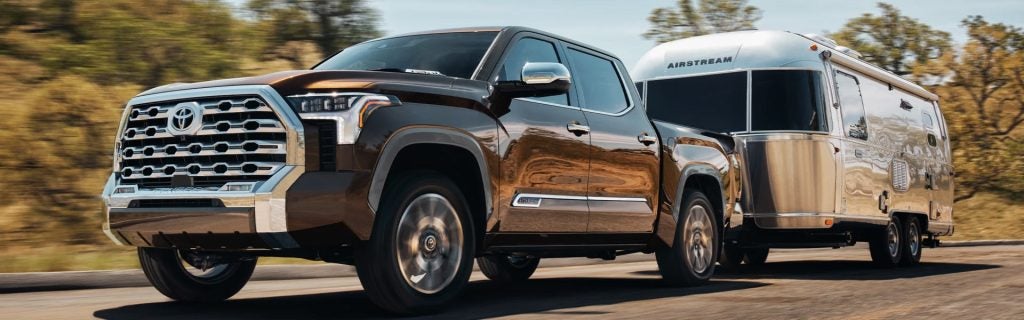 A brown Toyota Tundra truck with chrome accents pulls a silver Airstream trailer down a road.