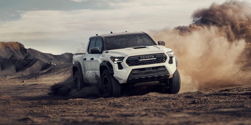 White Toyota pickup truck kicking up a large cloud of dust while driving through a rugged, arid landscape.