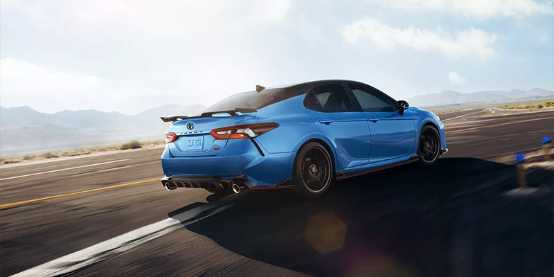 Rear three-quarter view of a metallic blue 2023 Toyota Camry TRD driving on an open highway.
