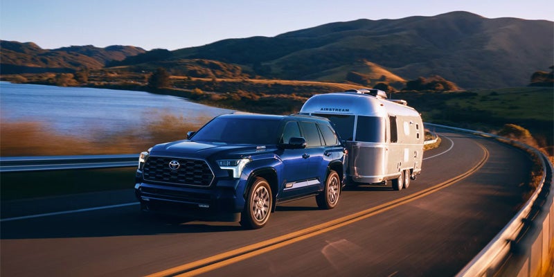 A dark blue SUV towing a silver Airstream trailer on a winding road next to a lake and mountains.