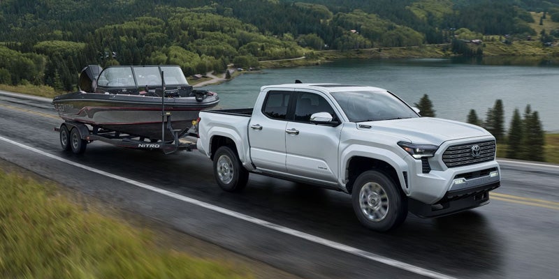Light gray pickup truck towing a black fishing boat on a trailer, driving on a road next to a lake and mountains.