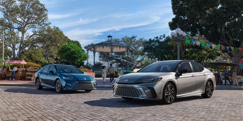 Two sleek Toyota sedans, a blue one and a silver one, parked on a cobblestone plaza with people and market stalls.