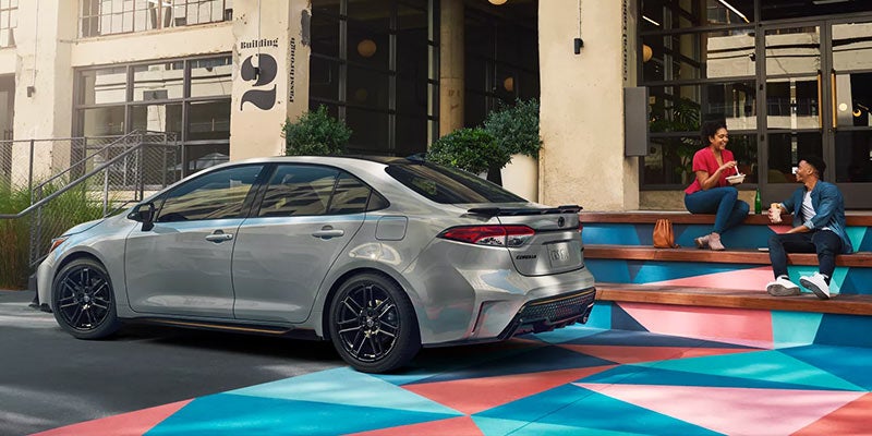 A silver Toyota Corolla parked on a colorful geometric-patterned pavement, with two people eating on steps in the background.