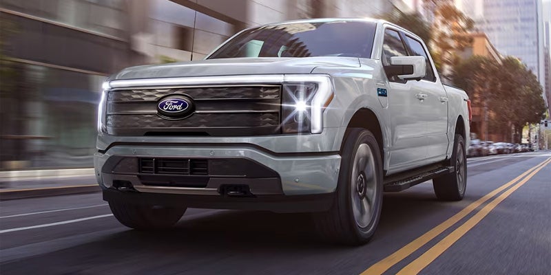 A light gray 2023 Ford F-150 pickup truck drives down a city street, headlights glowing.
