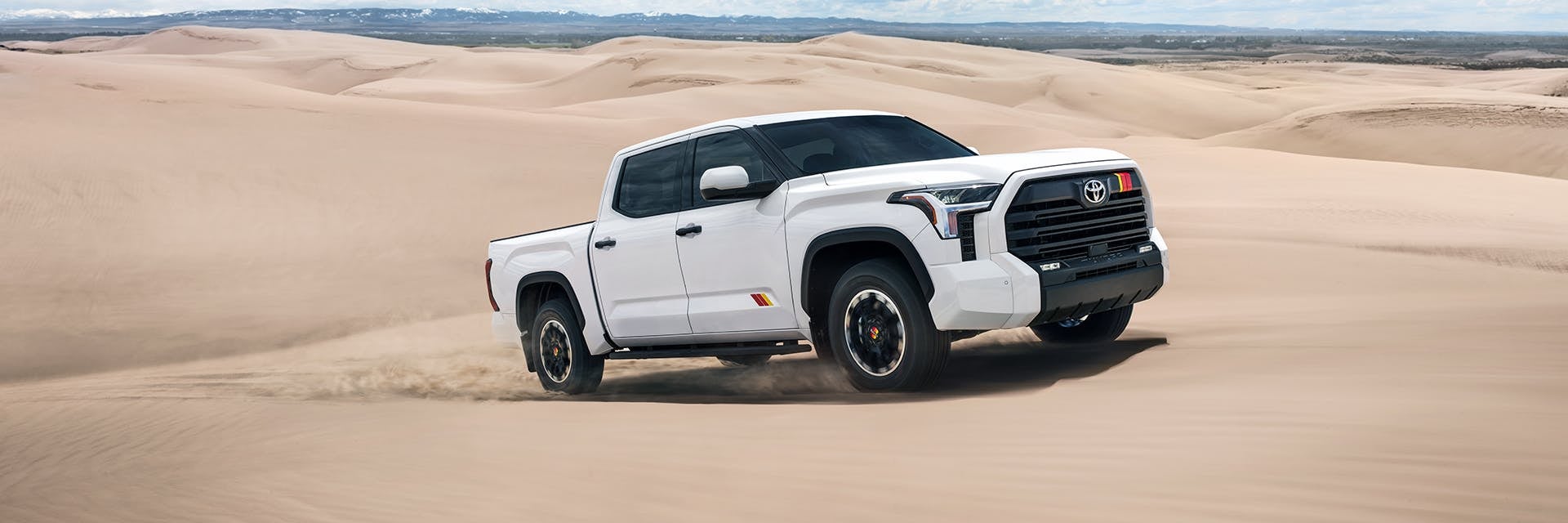 White Toyota Tundra pickup truck kicking up dust in a desert, showing multicolored racing stripes.
