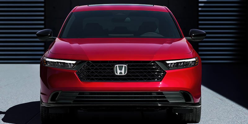 Front view of a new red Honda Accord sedan with its headlights on, parked in front of a dark building.