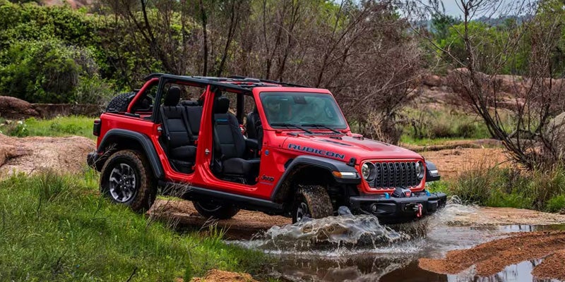 A doorless red Jeep Wrangler Rubicon driving through a shallow stream, splashing water as it moves.