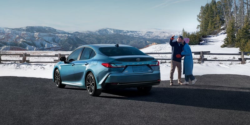 A blue Toyota Camry parked on a snowy overlook, with a couple taking a selfie next to it.