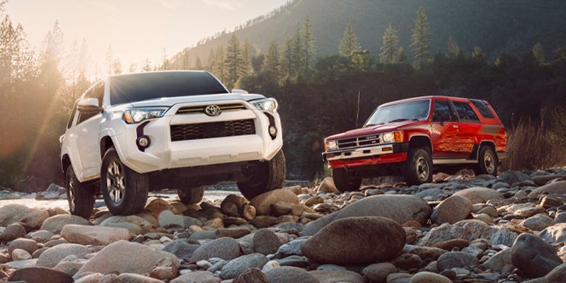 A white modern SUV and a red vintage SUV parked on a rocky river bed under a soft sunset.