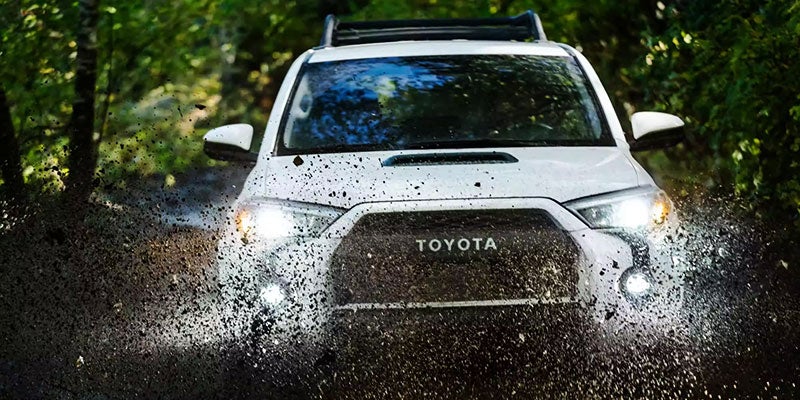 A white Toyota SUV drives through mud, splattering it over the front grille and headlights.