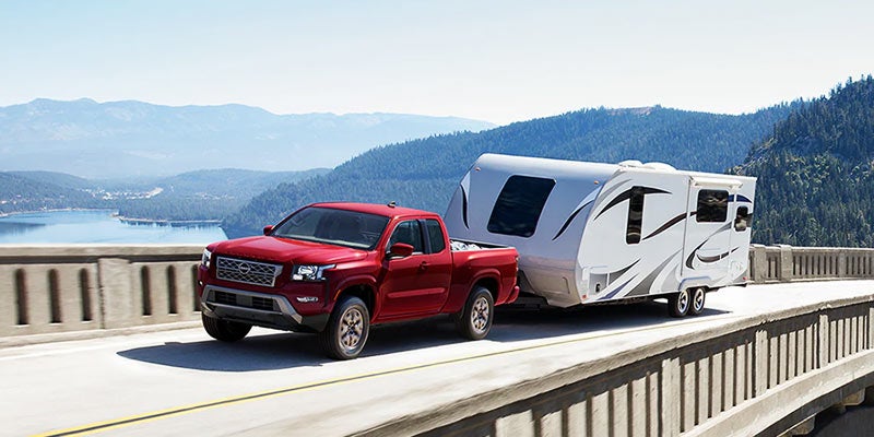 A red truck tows a white camper trailer along a bridge with a lake and mountains in the background.