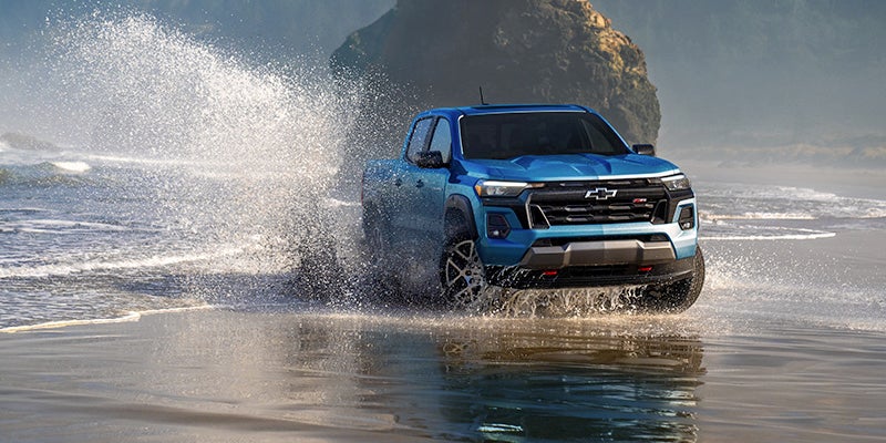 Blue Chevy Colorado ZR2 pickup truck drives through shallow water on a beach, splashing. Remote rocky island in background.