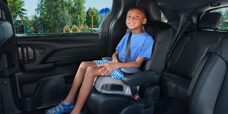 A young Black boy in a blue shirt sits in a booster seat in a car, looking out at a playground.