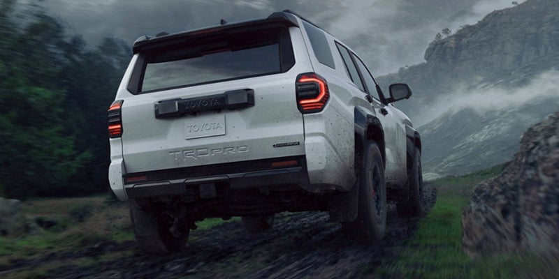 Rear view of a white Toyota TRD Pro SUV driving up a muddy incline on a misty, mountainous landscape.