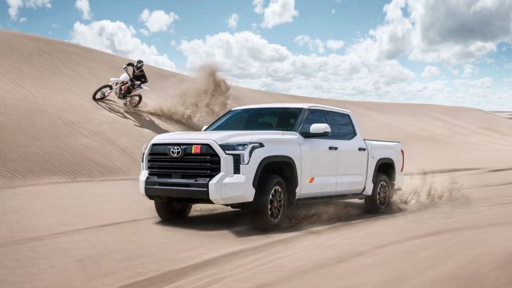 A white Toyota Tundra truck drives through sand dunes as a motocross rider goes up a steep dune in the background.
