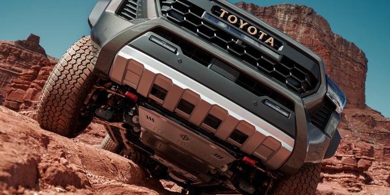 A brown Toyota truck's front end and underside driving up steep red rock terrain, with a mesa in the background.