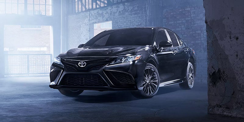A sleek, black Toyota Camry sedan with dark rims, parked in a dimly lit, dusty industrial building.