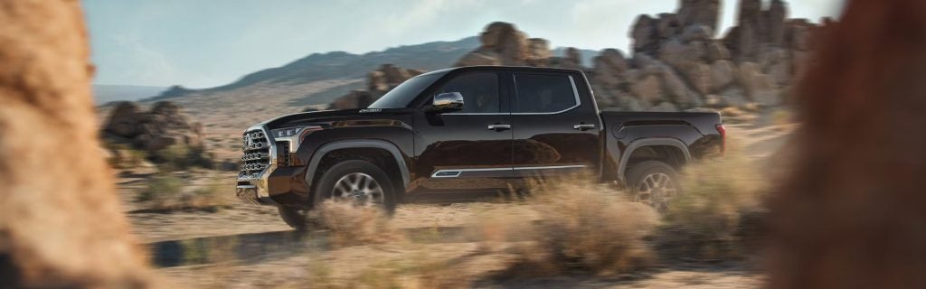 Dark brown pickup truck driving on a dirt road through a desert landscape with rocky outcrops.