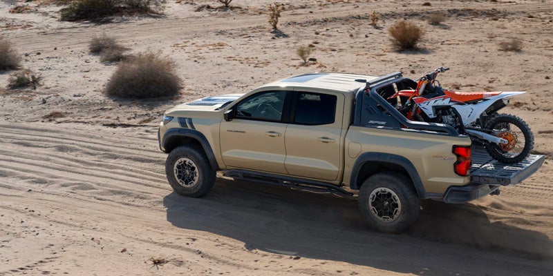 Khaki pickup truck with black trim and roll bar, a white and orange dirt bike in its bed, driving on sand.