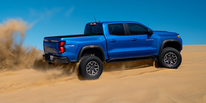 A blue Chevrolet Colorado pickup truck with large off-road tires kicking up sand in a desert dune.