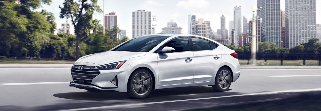 White 2019 Hyundai Elantra driving down a highway at the edge of a city.