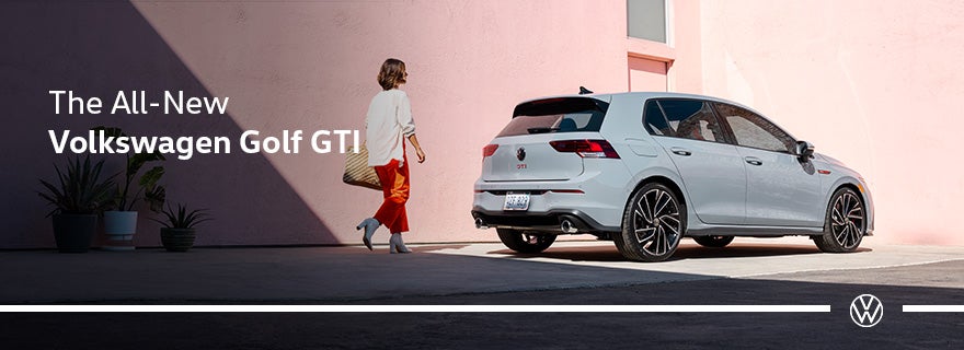 Person walking toward a white Volkswagen Golf GTI parked against a pink wall.