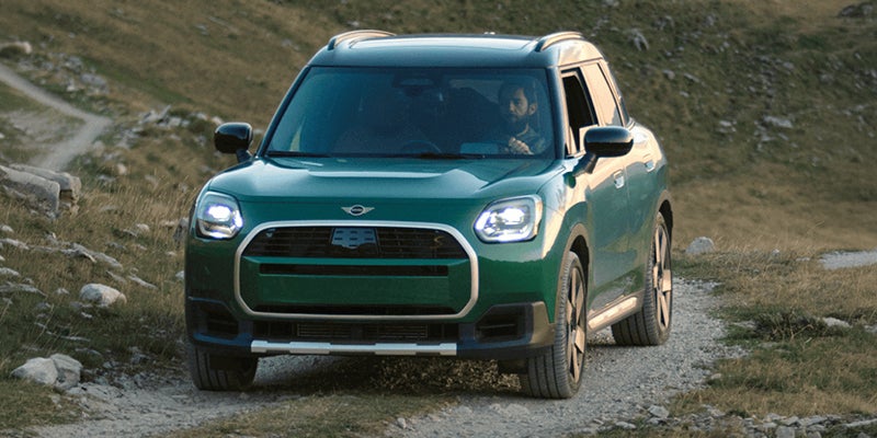 A green 2026 MINI Countryman John Cooper Works ALL4 drives along a rugged mountain trail with its LED headlights illuminated, showcasing sporty styling and all-terrain capability.