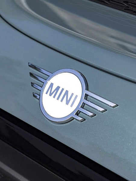 Close-up of a chrome MINI logo with wings on a car, against a green background.