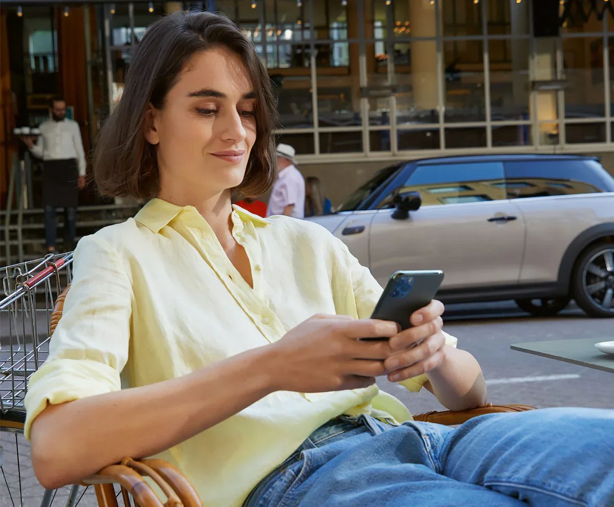 A young woman in a yellow shirt sits relaxed, using a smartphone outdoors, with a MINI parked nearby.