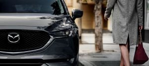 Close view of the front driver side of a gray 2020 Mazda CX-5 parked by a sidewalk with a woman wearing a long gray jacket standing beside it. | Mazda dealer in Ellisville, MO