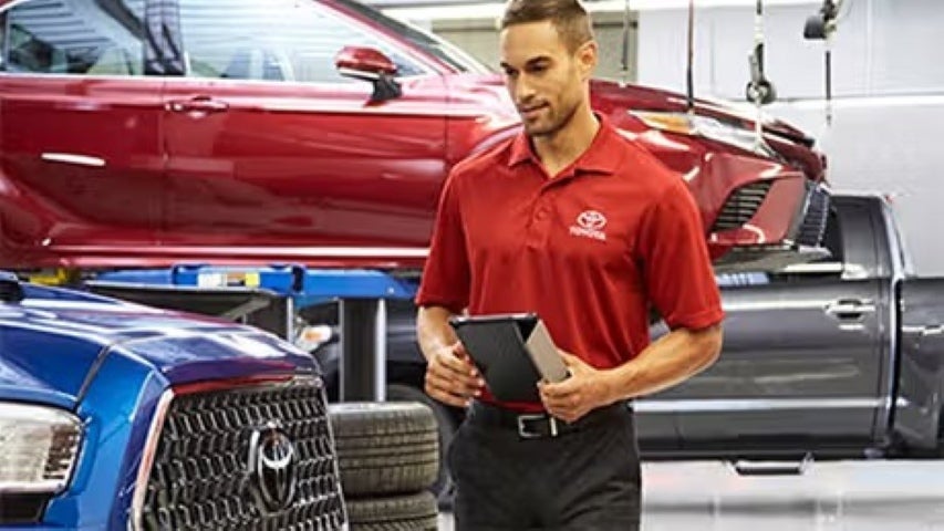 Toyota service advisor holding a tablet while inspecting a blue vehicle in a repair bay with cars on lifts.