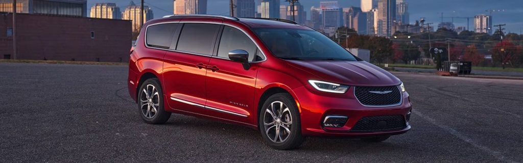 Red Chrysler Pacifica minivan parked in a lot with a city skyline at dusk.