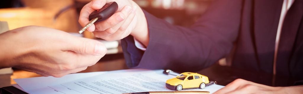 Car key being handed over during a vehicle purchase with paperwork and a toy car on the desk.