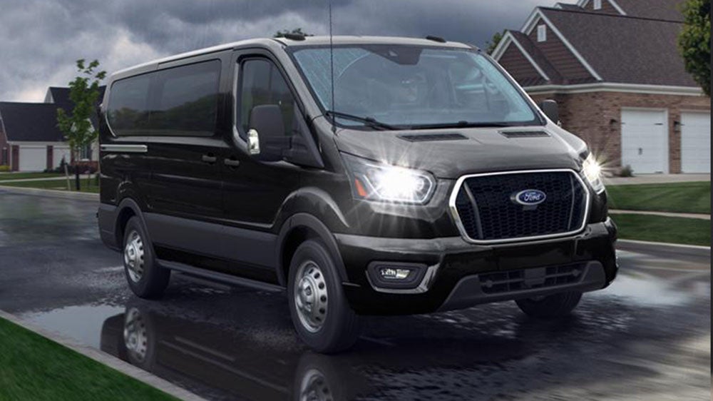 Exterior front-quarter view of a dark gray Ford Transit van driving on a residential street.