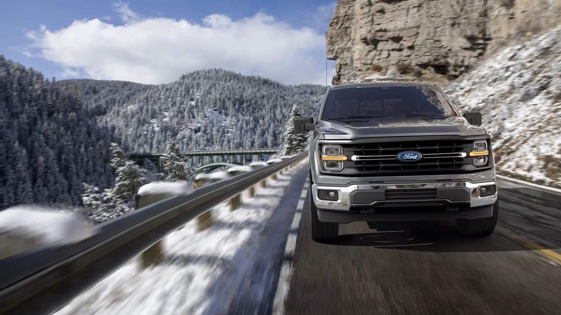 A silver Ford F-150 drives along a snowy mountain road beside a rocky cliff