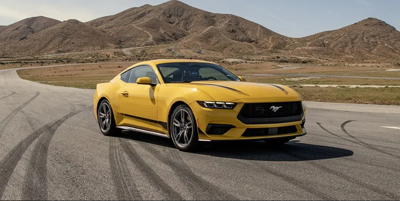 A yellow ford Mustang driving down a road surrounded by mountains.