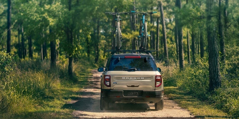 A tan 2024 Ford Bronco Sport driving down a dirt road on a sunny day with a beautiful wooded landscape on either side of the road