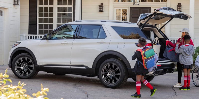 Two children prepare to load bags into the open trunk of a white SUV in a driveway.