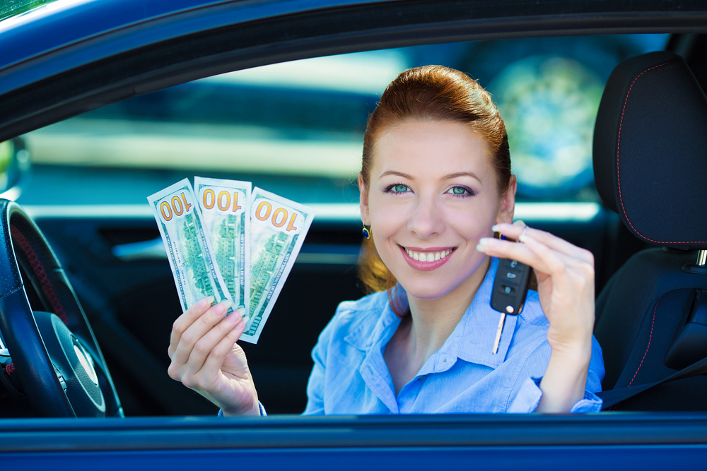 Smiling woman sitting in the driver’s seat of a car holding cash and a car key, suggesting savings or a successful car purchase, with a bright and modern interior background.
