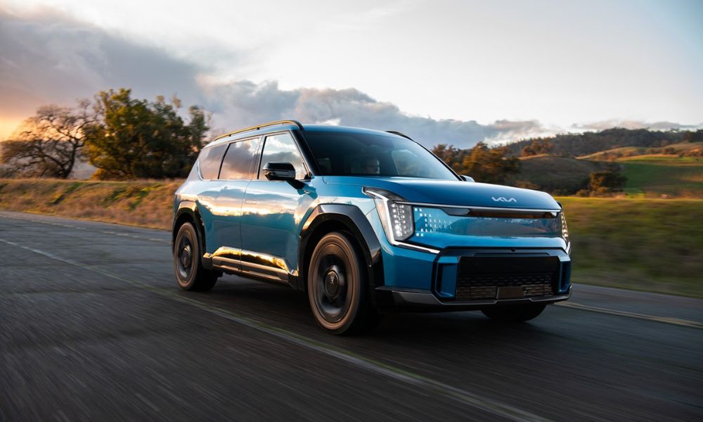 A blue Kia EV9 electric SUV driving on a scenic road during sunset, showcasing its bold design, modern headlights, and aerodynamic build.