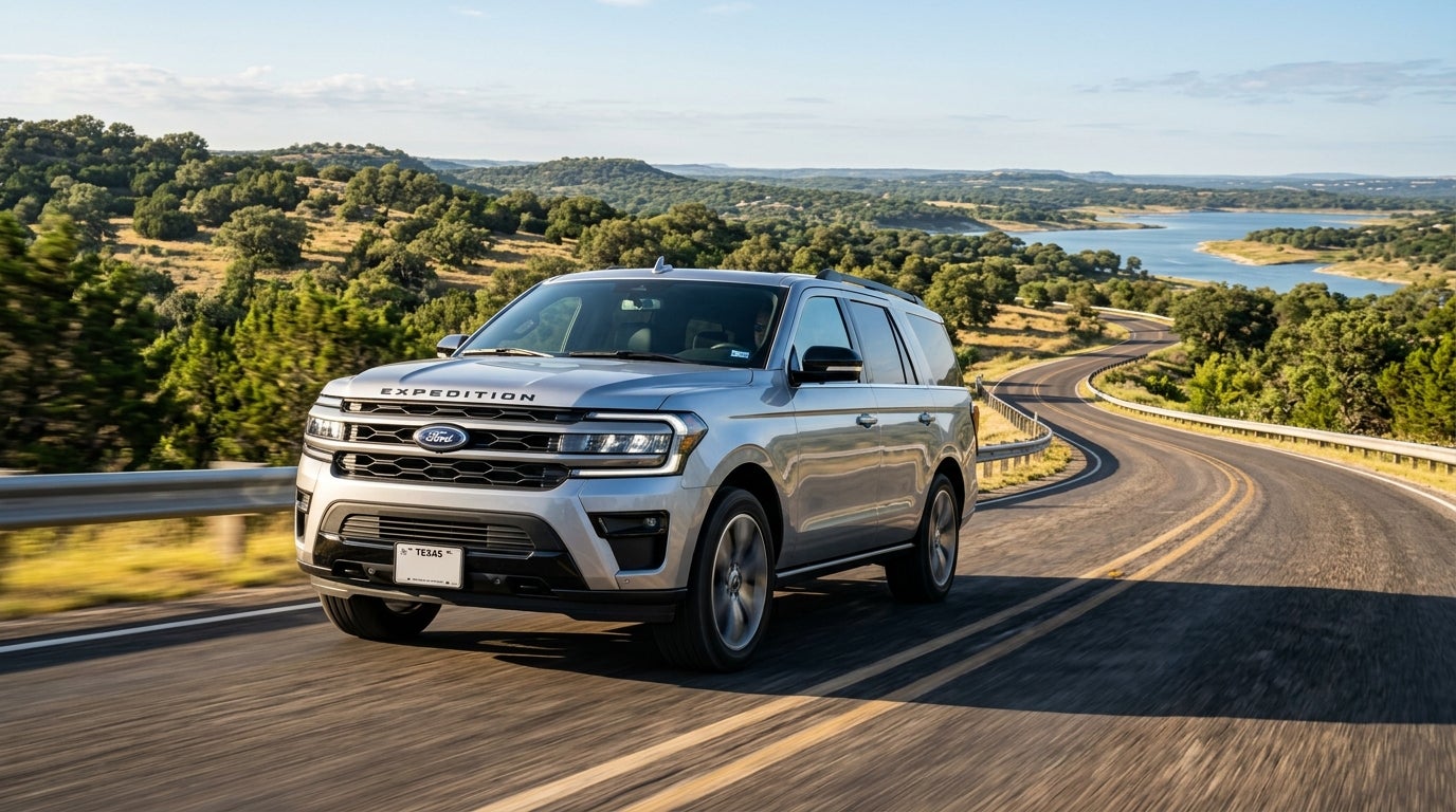 Silver Ford Expedition driving on a winding road through a hilly landscape with a blue lake in the background.