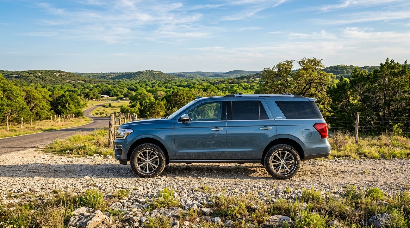 Profile view of a blue SUV parked on a gravel path overlooking a lush green valley with a winding road.