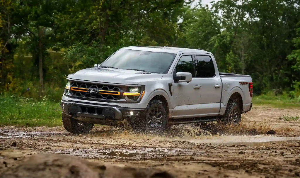 A silver Ford F-150 pickup truck driving through a muddy area with greenery in the background.