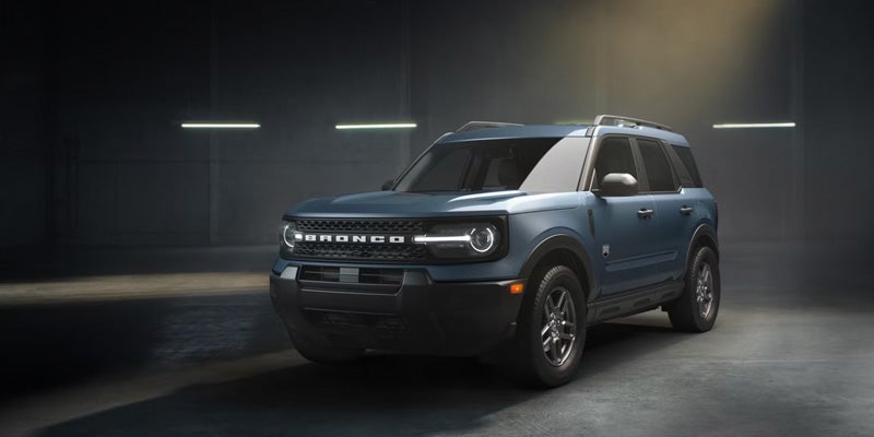 A 2026 Ford Bronco Sport parked in a dimly lit space, highlighting its rugged design and distinctive front grille with "BRONCO" lettering.