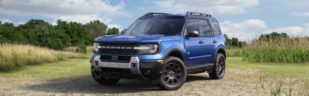 A blue 2026 Ford Bronco Sport SUV parked on a gravel surface with grassy fields and trees in the background.