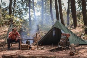 Man sitting by smoky campfire enjoying a drink while on a camping ground with his tent set up.