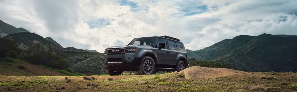 A grey Toyota Land Cruiser parked on a grassy hilltop with mountains in the background.