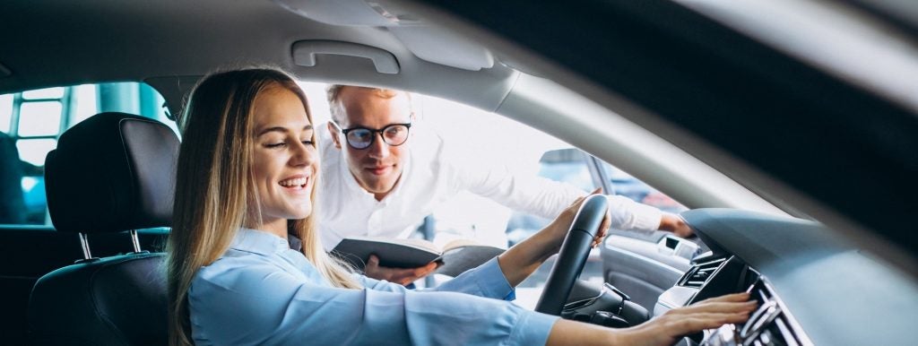 A car salesman assisting a woman sitting in the driver's seat.