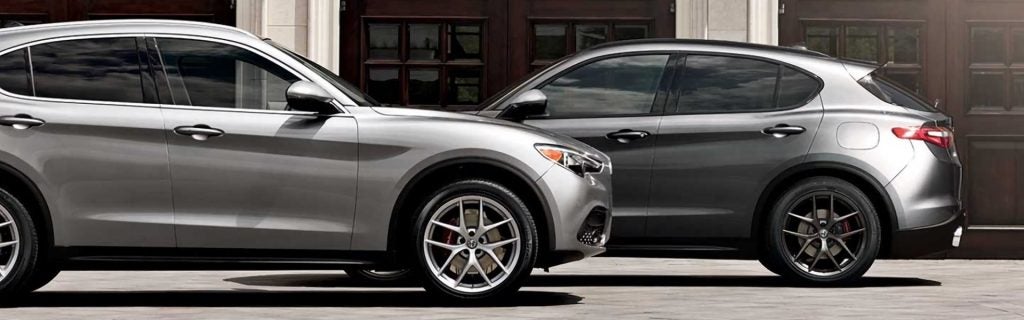 A silver Alfa Romeo Stelvio SUV parked in front of a dark gray Alfa Romeo SUV, showcasing their side profiles and sporty alloy wheels against a background of dark wooden doors.