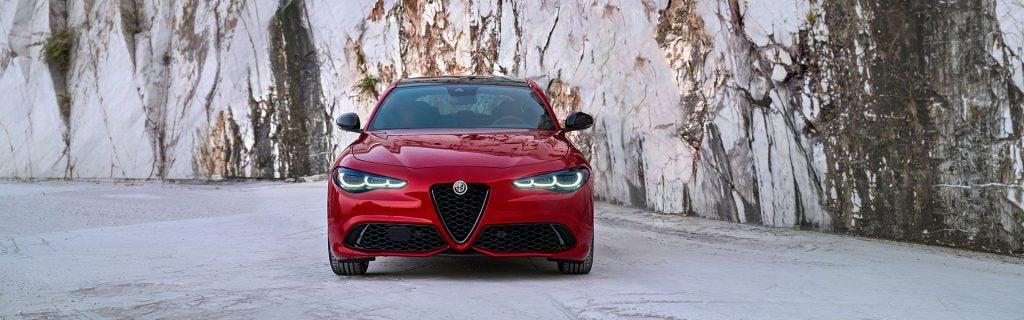 Front view of a red sports sedan parked on a concrete surface, framed by tall, textured white stone walls, with its LED headlights on and distinctive triangular grille centered.